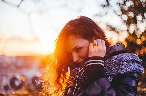 Nainen seisoo ilta-auringossa ja kuuntelee korvanapeista musiikkia.